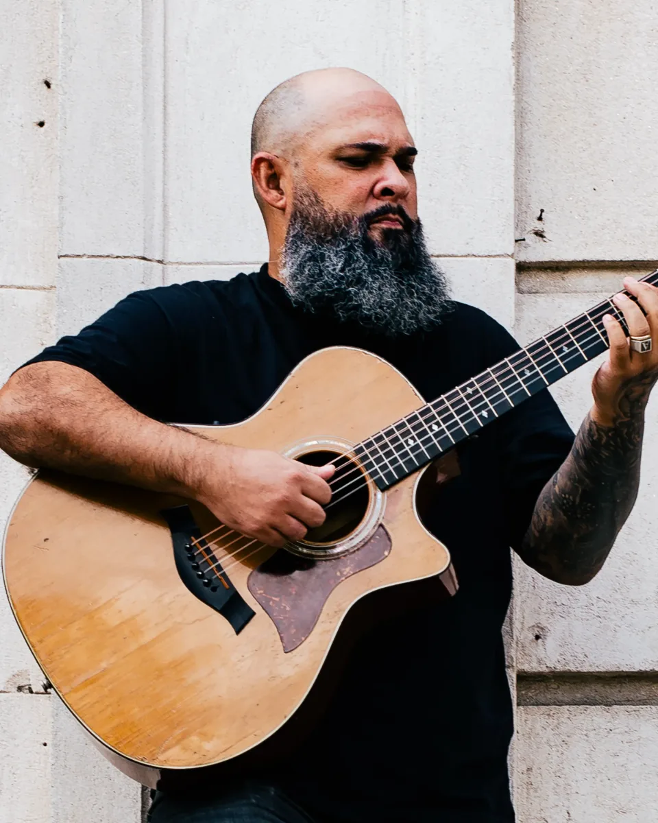 Vitor Teixeira tocando violão junto a uma parede de pedra