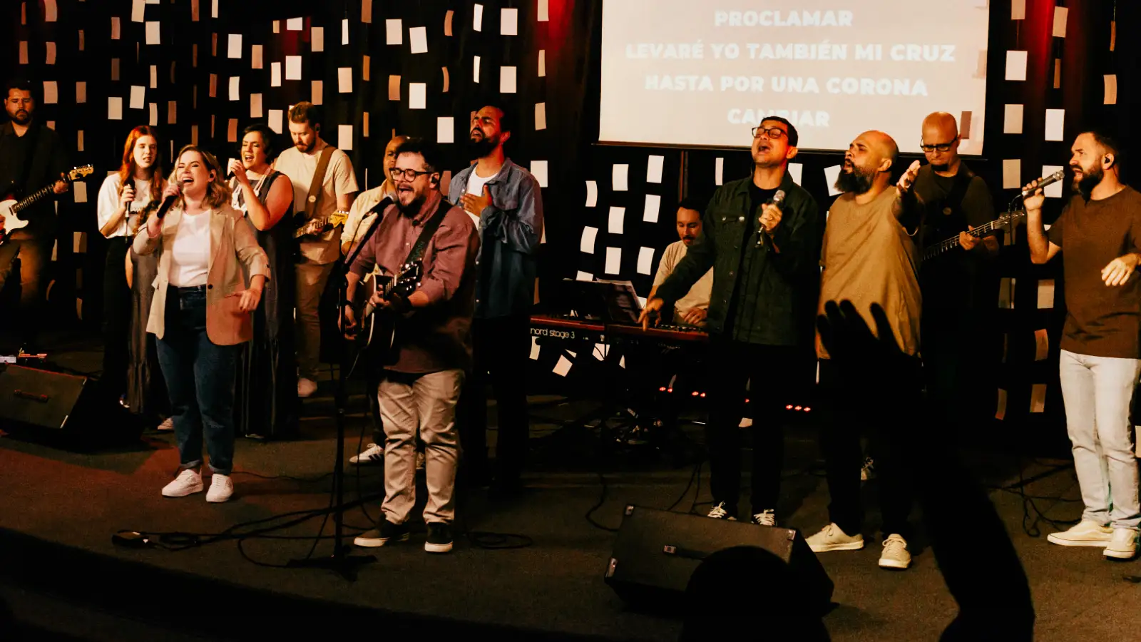 Carlinhos Falsetti canta ao violão ao lado da banda e dos vocalistas durante a gravação do álbum Alabanza, com a letra de «Rude Cruz» em espanhol projetada ao fundo
