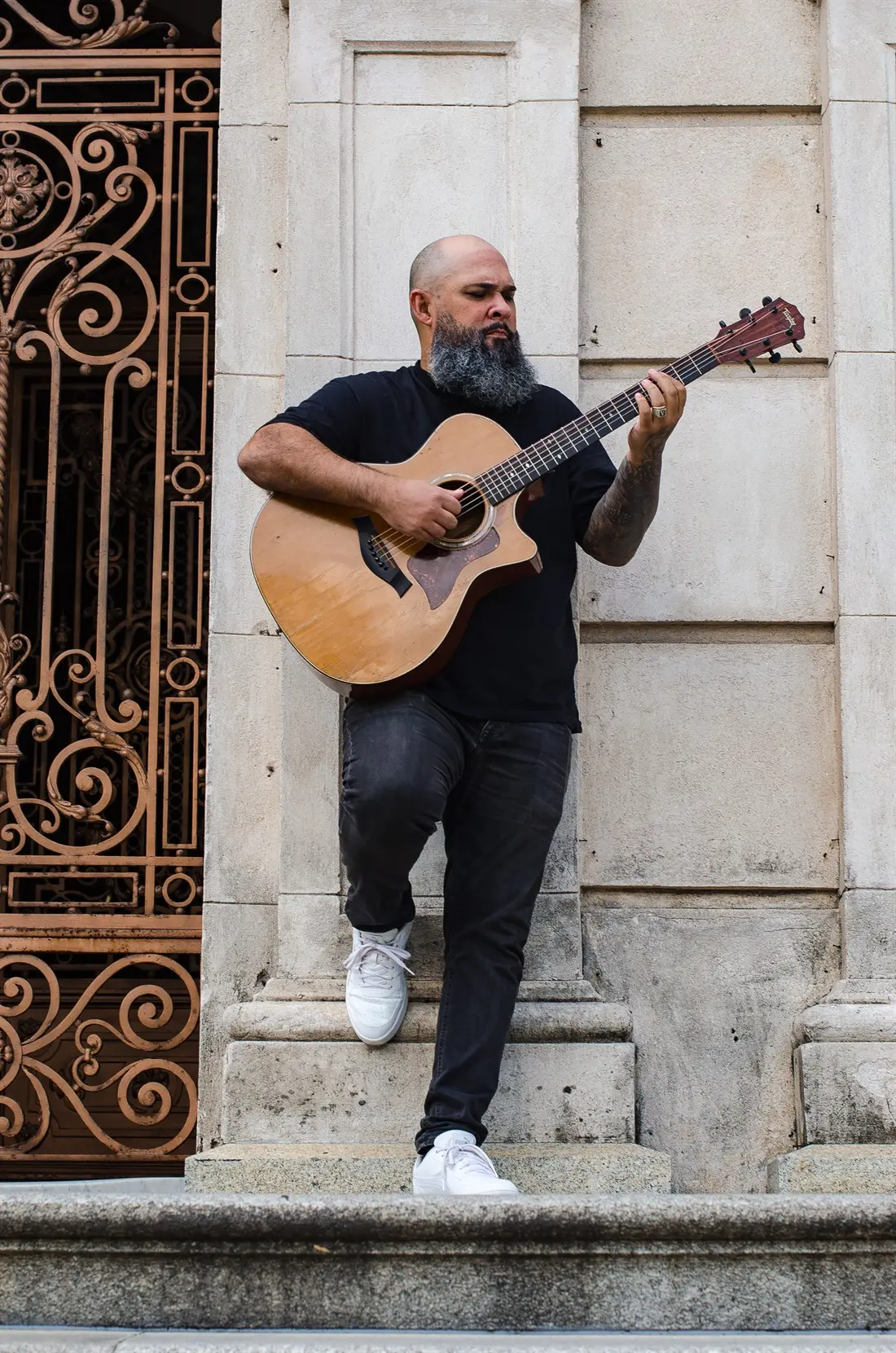 Vitor Teixeira tocando violão em pé, diante de uma fachada de pedra e de um portão de ferro
